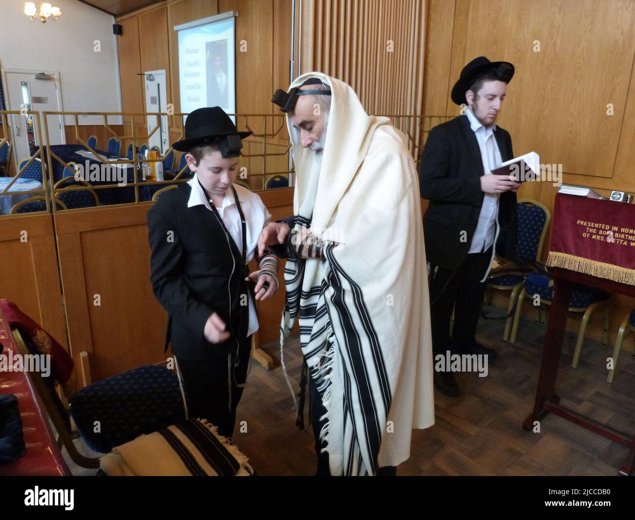 Wenige Wochen vor seiner Barmitzvah-Zeremonie und Feier wird Menachem Mendel Pink von seinem Vater Rabbi Yehuda Pink von der Solihull and District Hebrew Congregation in den West Midlands zum ersten Mal beim Anziehen von Tephillim unterstützt und von seinem Großvater, Herrn Phaivish Pink, beobachtet. Der Schulleiter des Manchester Jewish Grammar School, bei einem Gottesdienst am Sonntagmorgen, gefolgt von einem Festfrühstück für seine Familie und andere Gläubige aus den Chasidic-Gemeinden in Lubavitch in den West Midlands sowie der Gemeinde der Synagoge. Stockfoto