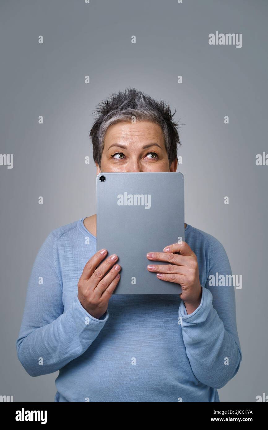 Grauhaarige reife Frau Angst verstecken Gesicht hinter digitalen Tablet-Posting negative, toxische Kommentar online, Social Media. Hübsche Frau mit Tablet in blauem Hemd isoliert auf weißem Hintergrund. Stockfoto