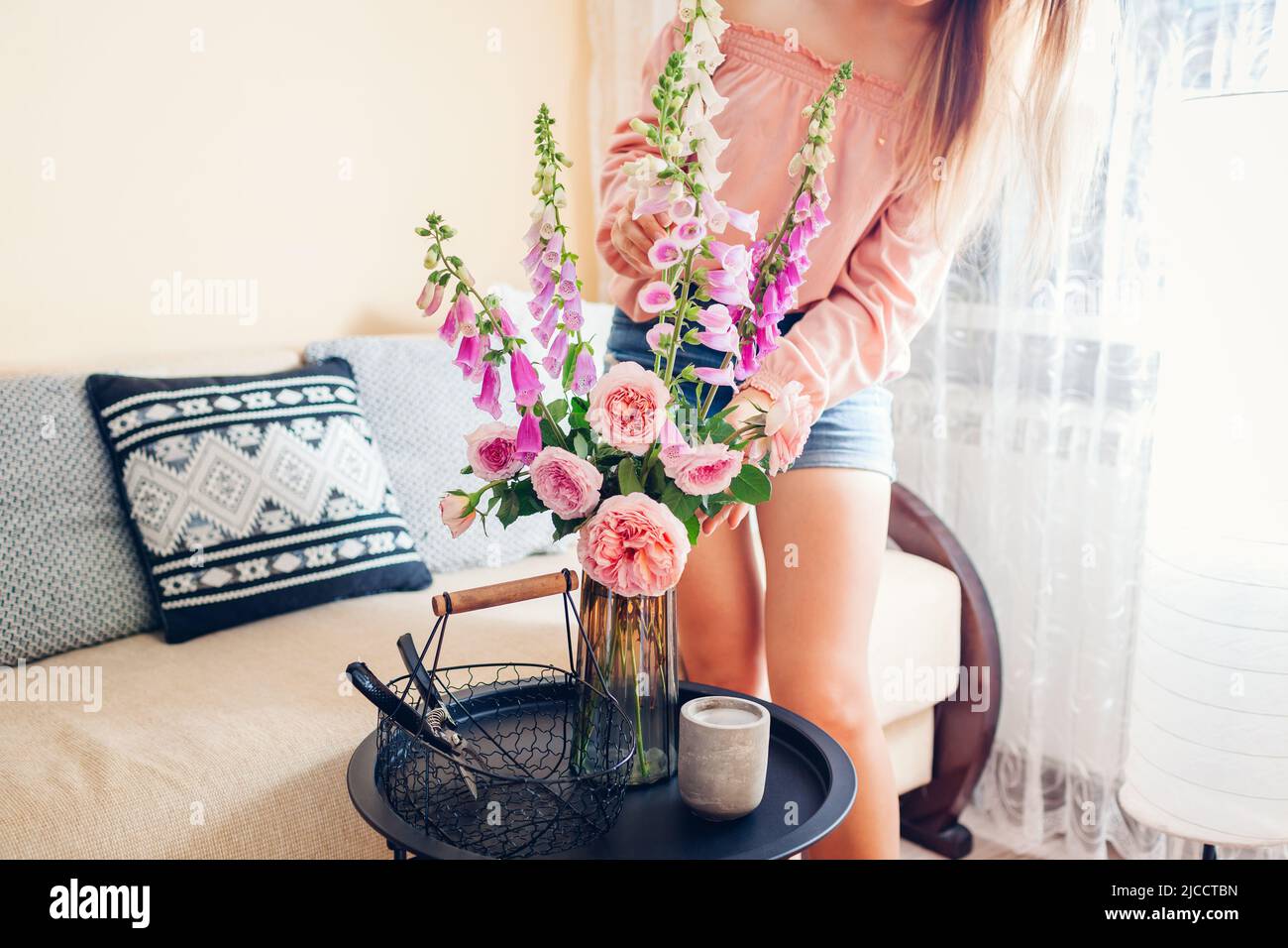 Frau legt Strauß Rosen und Füchshandschuhen Blumen in Korb in Vase zu Hause gepflückt. Einrichtung und Einrichtung im Sommer. Frische rosa lila orange Blüten von aust Stockfoto