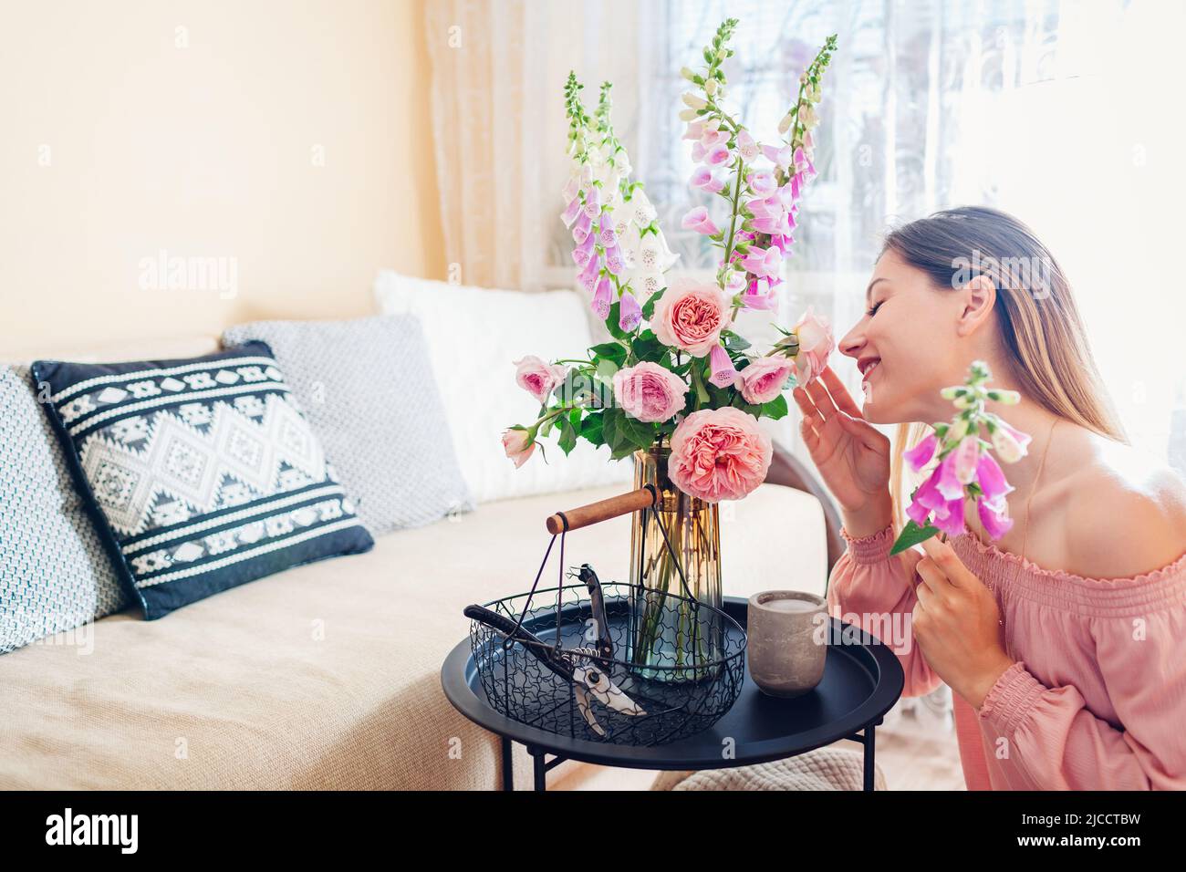 Frau riecht Blumen und arrangiert Strauß mit Rosen und Füchshandschuhen zu Hause. Frische Blüten in Vase auf den Tisch gelegt. Einrichtung und Einrichtung im Sommer Stockfoto