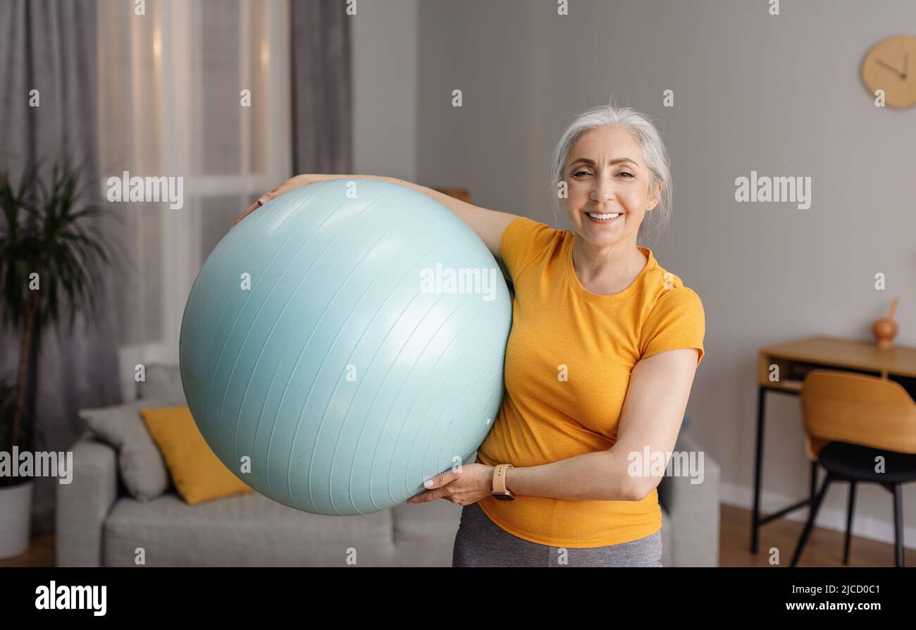 Porträt einer positiven älteren Frau, die den Fitnessball hält und die Kamera anlächelt und zu Hause im Wohnzimmer trainiert Stockfoto