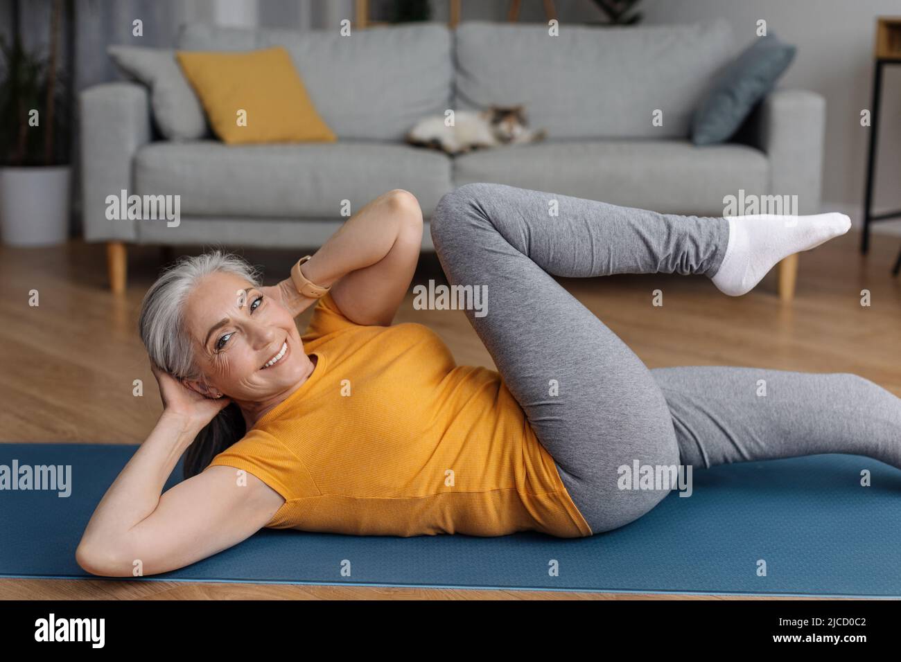 Aktive ältere Frau, die zu Hause auf einer Yogamatte ABS-Übungen macht und die Kamera anlächelt und im Wohnzimmer trainiert Stockfoto