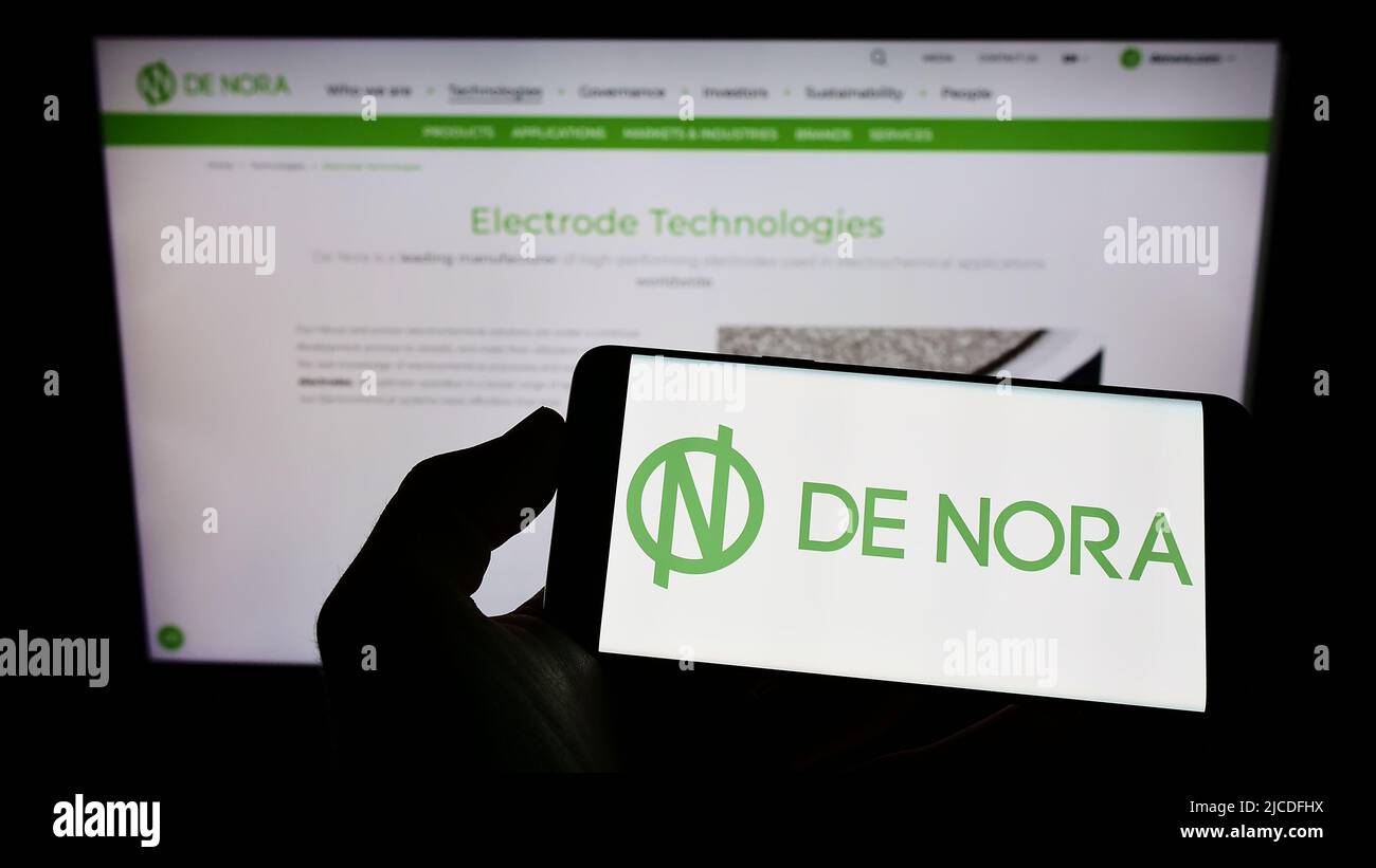 Person, die Mobiltelefon mit dem Logo der italienischen Firma Industrie De Nora S.p.A. auf dem Bildschirm vor der Business-Webseite hält. Konzentrieren Sie sich auf die Telefonanzeige. Stockfoto