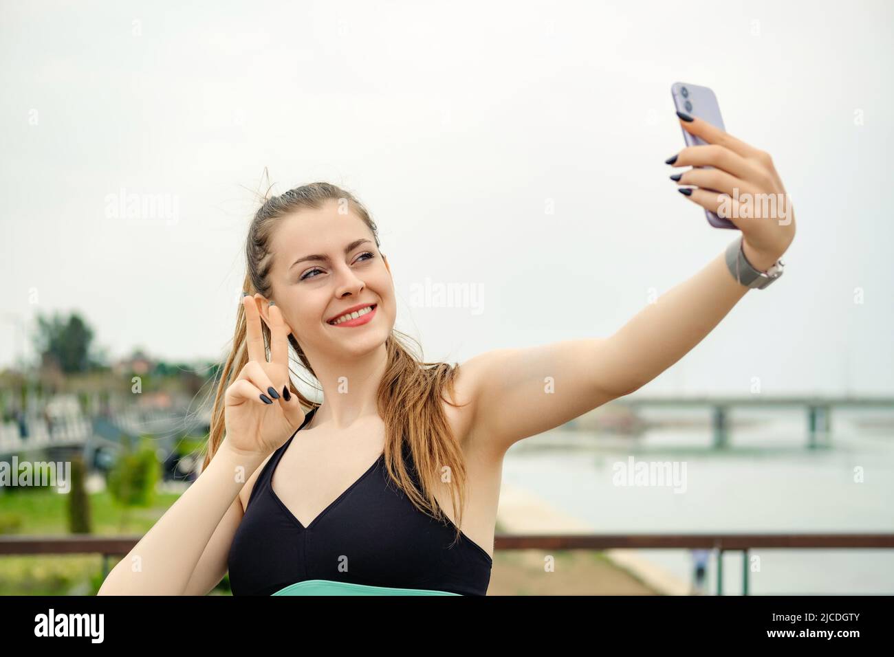 Junge Frau, die selbstbewusst lächelt, trägt einen Sport-BH, der im Stadtpark steht, im Freien lächelt und mit einem glücklichen Gesicht zuwinkt, wenn das Smartphone das Siegeszeichen tut Stockfoto