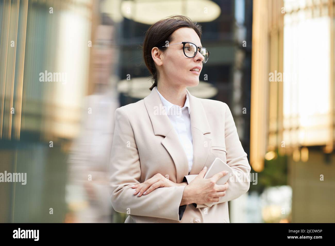 Eine zielgerichtete, attraktive Geschäftsfrau mittleren Alters in stilvoller Jacke, die in der Lobby steht und das Smartphone hält, während sie auf einen Kollegen wartet Stockfoto