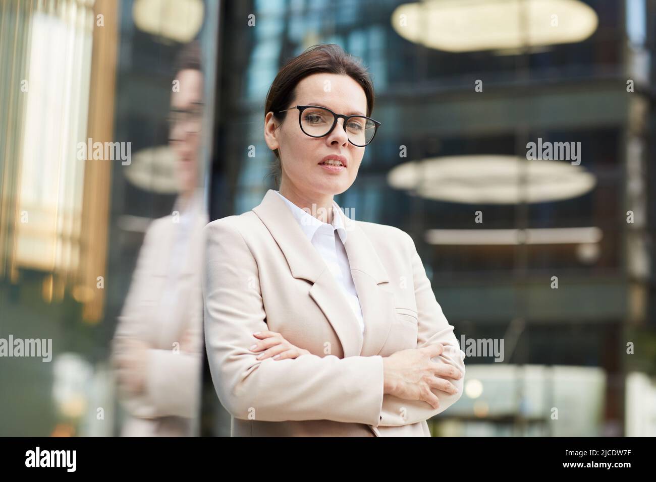 Content-Porträt eine selbstbewusste Dame mittleren Alters in stilvoller Jacke und Brillen, die die Arme auf der Brust kreuzt, während sie in der Lobby des Business Centers steht Stockfoto