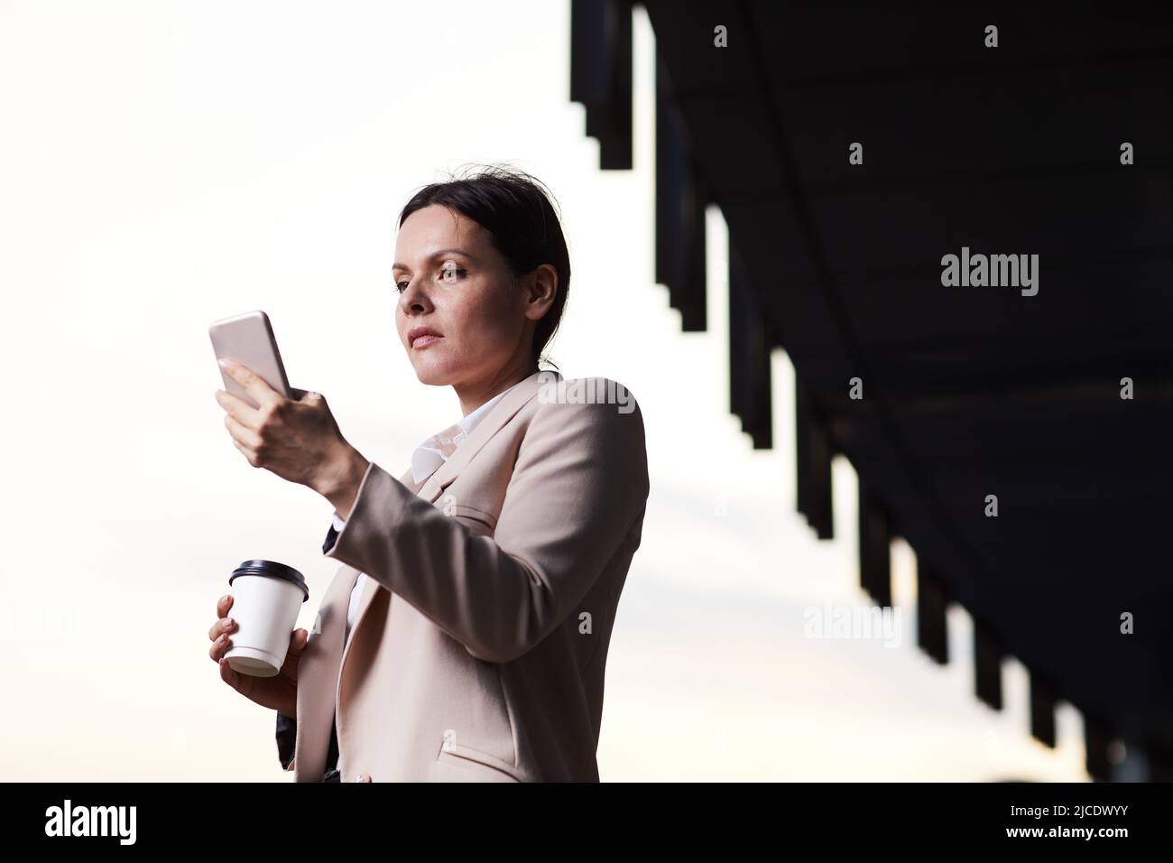 Seriöse Geschäftsfrau mittleren Alters in stilvoller Jacke, die im Freien steht und das Telefon nutzt, während sie das Internet-Signal empfängt Stockfoto