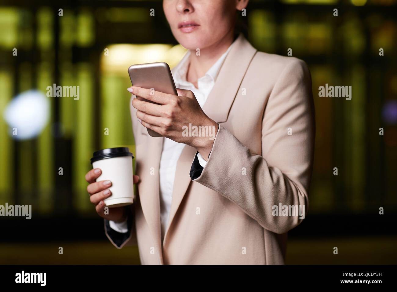 Nahaufnahme einer durchdachten Geschäftsfrau in stilvoller Jacke, die im dunklen Büro steht und Kaffee trinkt, während sie die Planner-App auf dem Smartphone überprüft Stockfoto