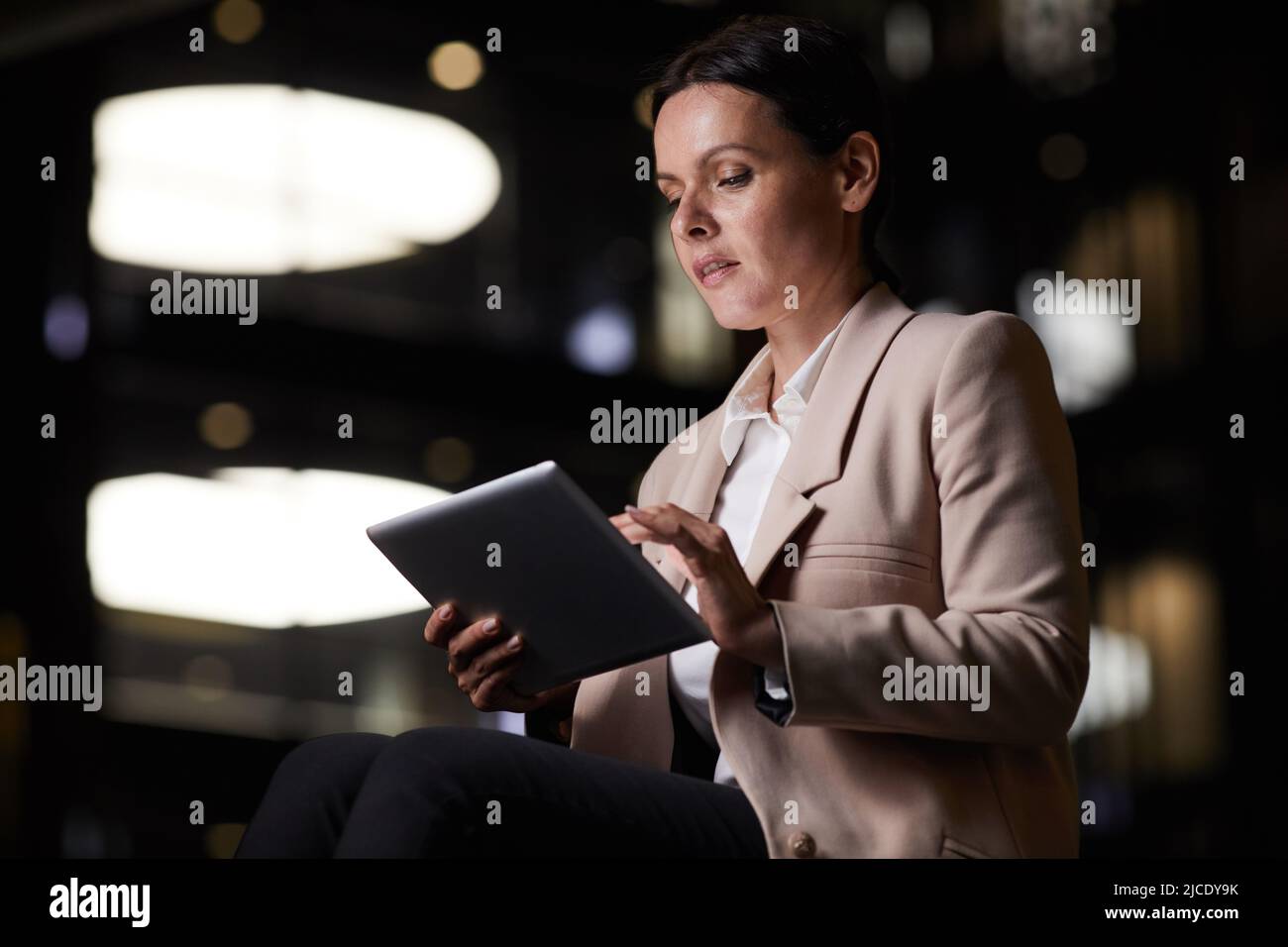 Eine durchdachte Geschäftsfrau mittleren Alters in stilvollem Outfit, die ein Tablet verwendet, während sie während der Arbeit in der Nacht Statistiken aus den sozialen Medien aufnahm Stockfoto