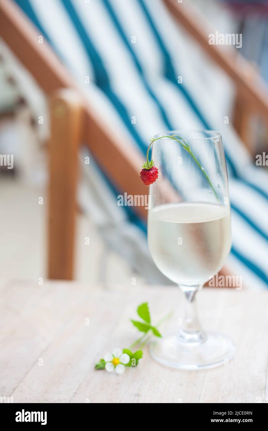 Ein Glas Weißwein mit Erdbeere auf dem Hintergrund eines Liegestuhls am Strand. Sommerzeit. Sommerliche Stimmung. Stockfoto