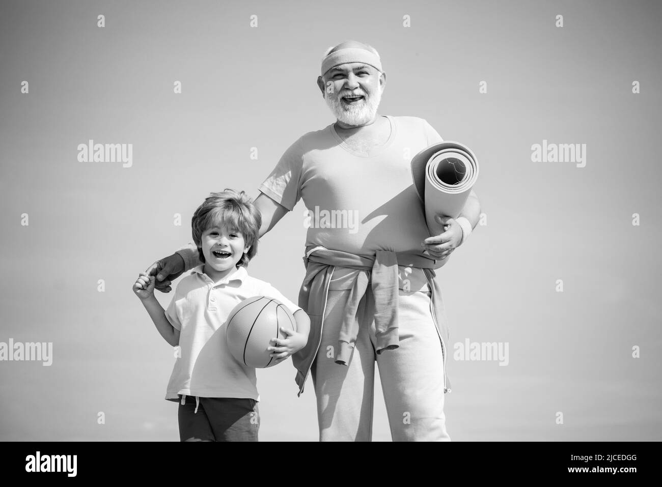 Großvater und Enkel mit Basketballball und Yogamatte in den Händen. Älterer Mann und niedlicher kleiner Junge trainieren auf blauem Himmel Hintergrund - isoliert. Stockfoto