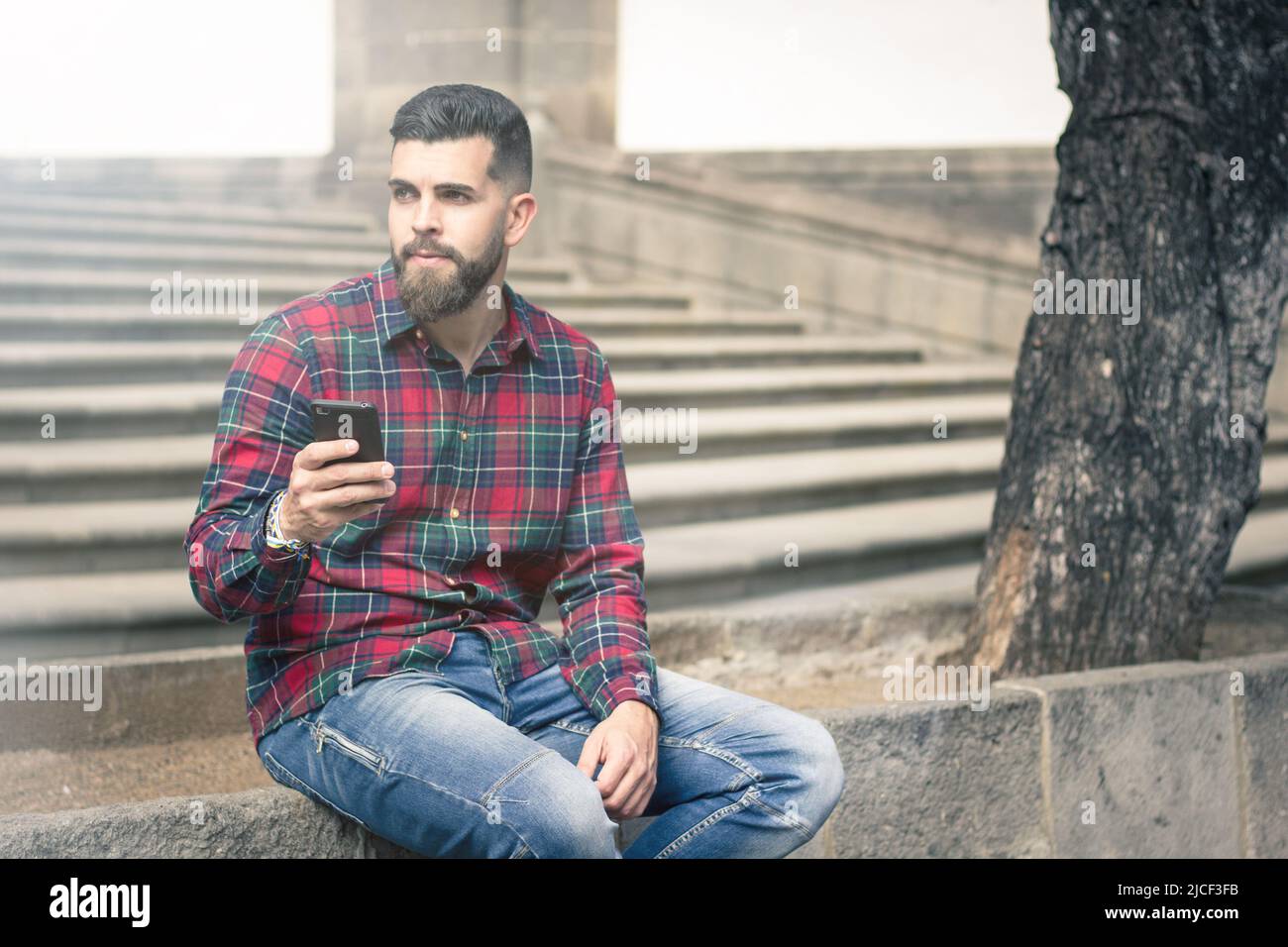 Junger Hipster mit Zellular, der in der Altstadt an einer Steinmauer an einem Baum sitzt. Nachdenklicher, gutaussehender Mann, der Smartphone auf der Straße hält. Lichteffekt Stockfoto