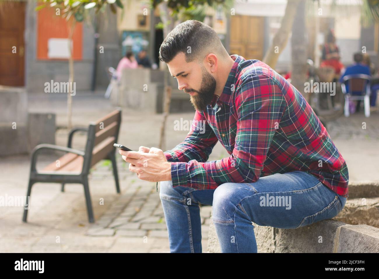 Hübscher Mann mit Vollbart und rotem Flanellhemd mit roten Schachbrettmuster und Smartphone auf dem Altstadtplatz von Las Palmas, Spanien. Junge Hipster mit Handy Stockfoto