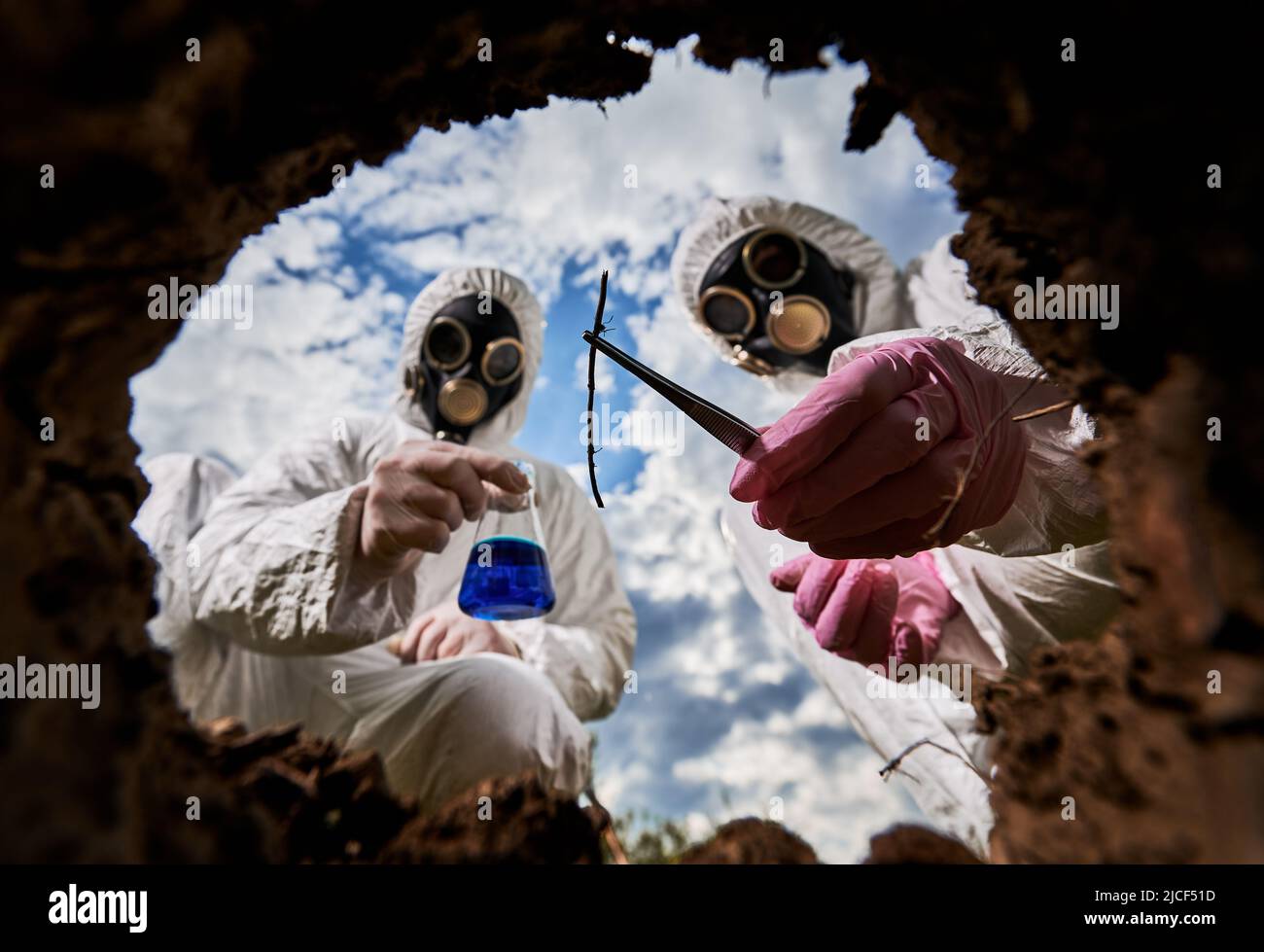 Wissenschaftler in Gasmasken und Schutzoveralls, die Reagenzglas mit blauer Flüssigkeit und Probe halten während eines gemeinsamen Experiments im radioaktiven Bereich untersuchen Fachleute die Verschmutzung. Blick von der Erdgrube. Stockfoto