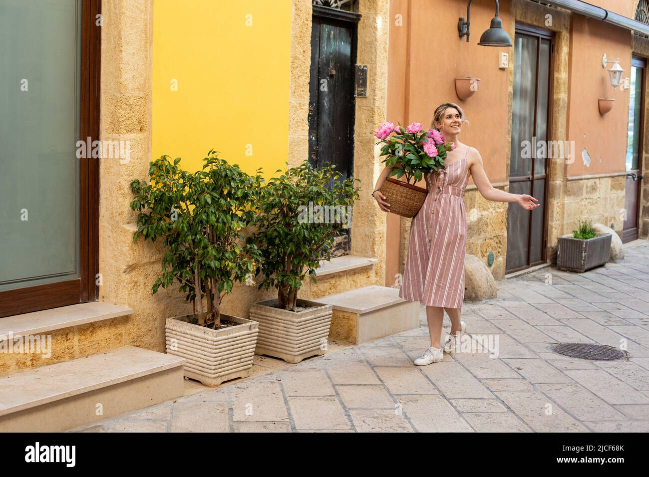 Junge lächelnde Frau im Sommer rosa gestreiften Kleid zu Fuß in der Nähe von Olivenbäumen, hält Topf mit rosa Pfingstrosen Blumen. Stockfoto
