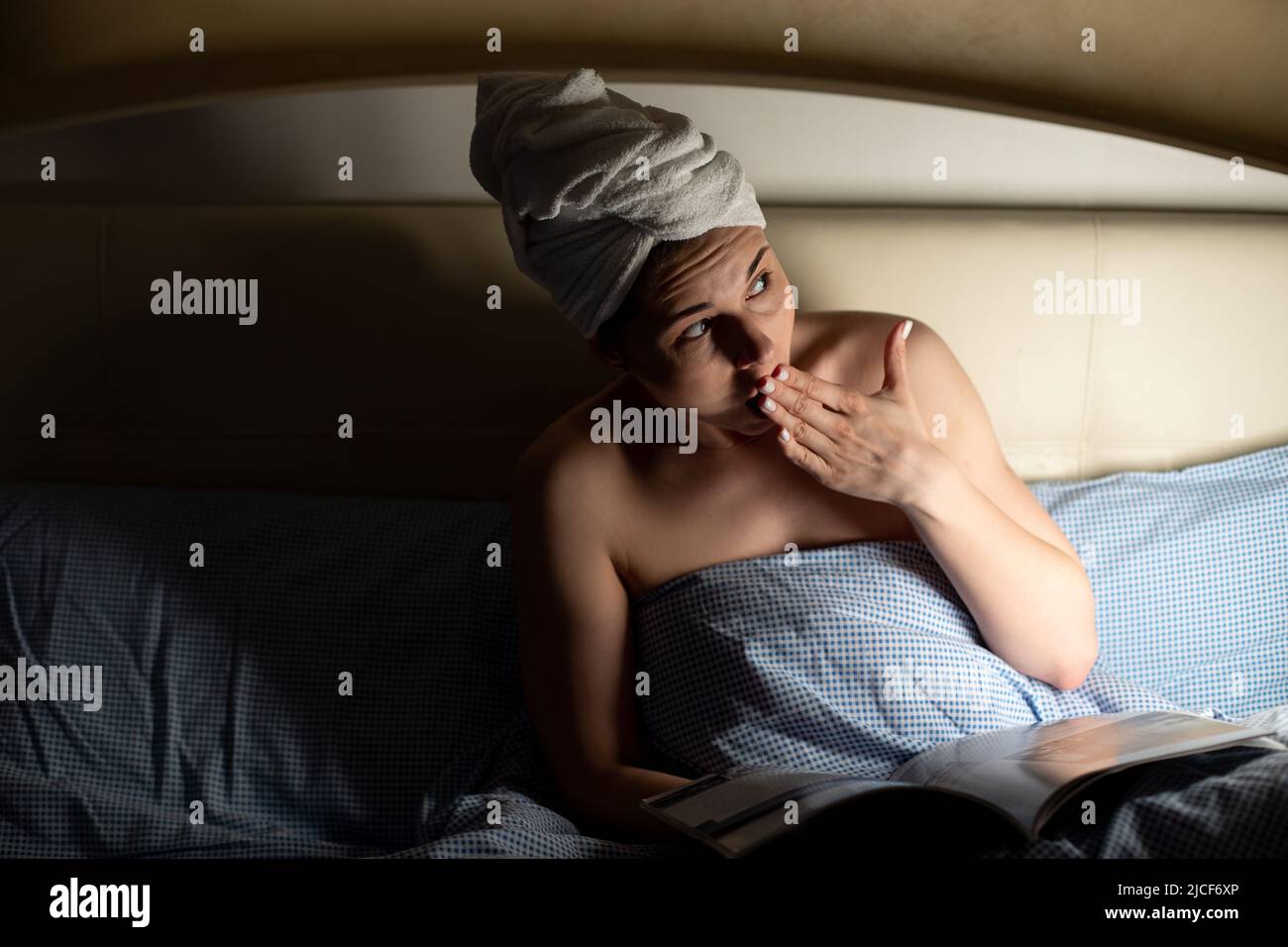 Porträt einer jungen, ängstlich verängstigten Frau, die auf dem Bett liegt, bedeckt mit einer blauen Decke, zur Seite schaut und den Mund mit der Hand bedeckt. Stockfoto