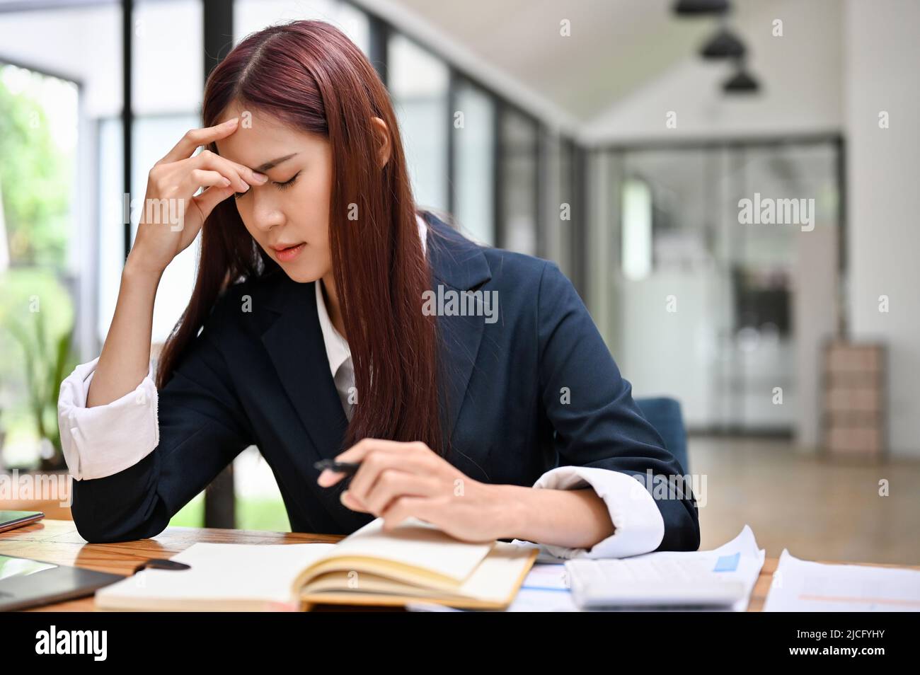 Gestresste junge asiatische Geschäftsfrau, die sich müde, erschöpft und aufgeregt über ihr Arbeitsergebnis fühlt. Seriöse Geschäftsfrau sitzt auf ihrem Schreibtisch. Stockfoto