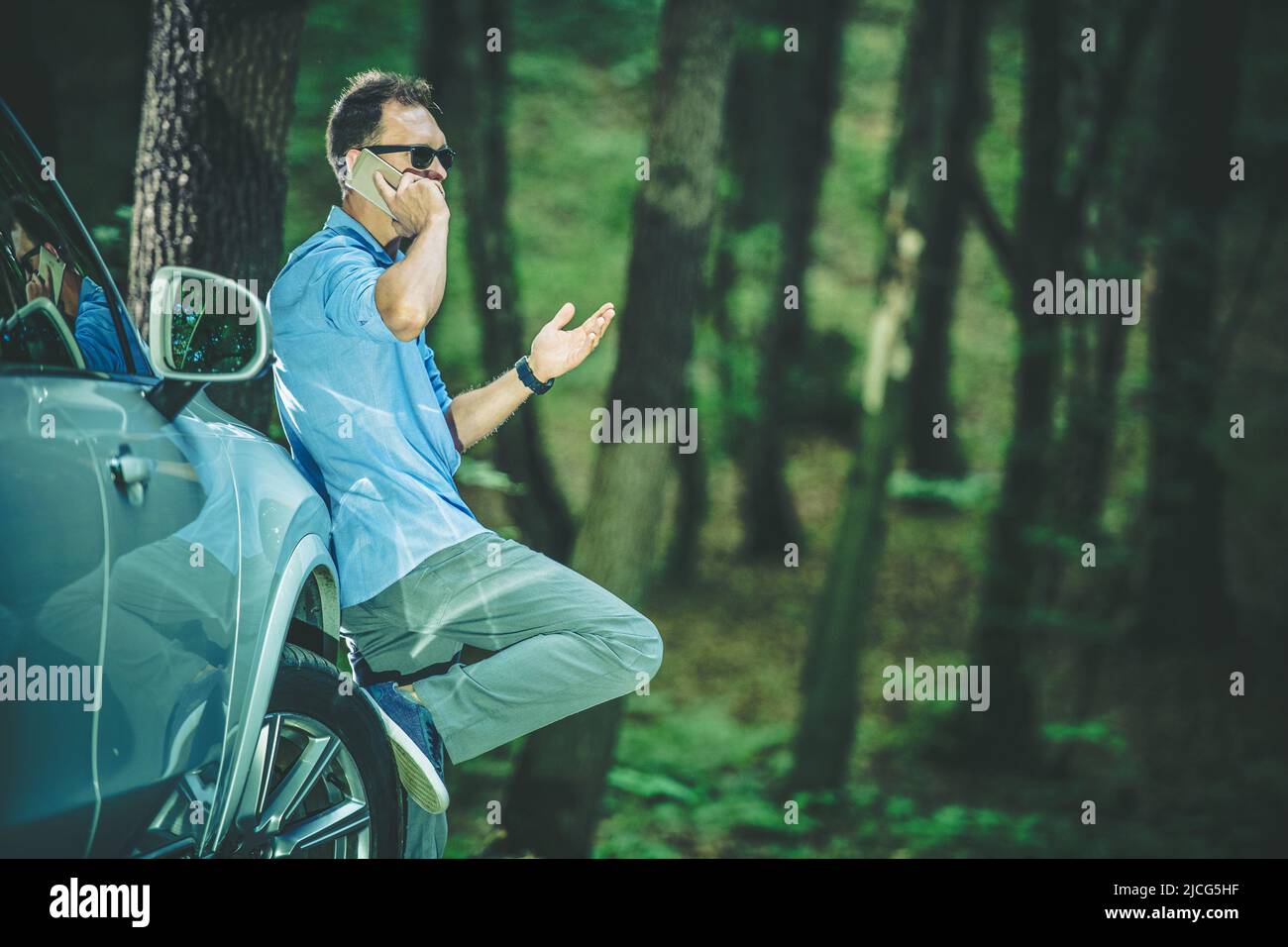 Kaukasischer Mann, der ein angespanntes Telefongespräch führt, nervös gestikuliert und seinen Fuß gegen das Rad seines Silberautos schief. Deep Forest im Stockfoto