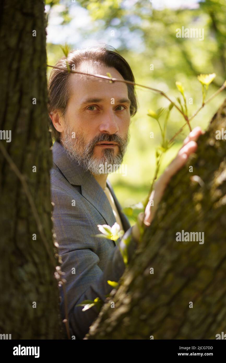 Reifer grauer Bart Geschäftsmann mit halblangen Haaren in lässiger grauer Jacke, die durch Baumstämme schaut, die sich auf einen stützen. Leben nach 40 Jahren Konzept, Probleme und Depressionen. Krise des mittleren Alters. Stockfoto