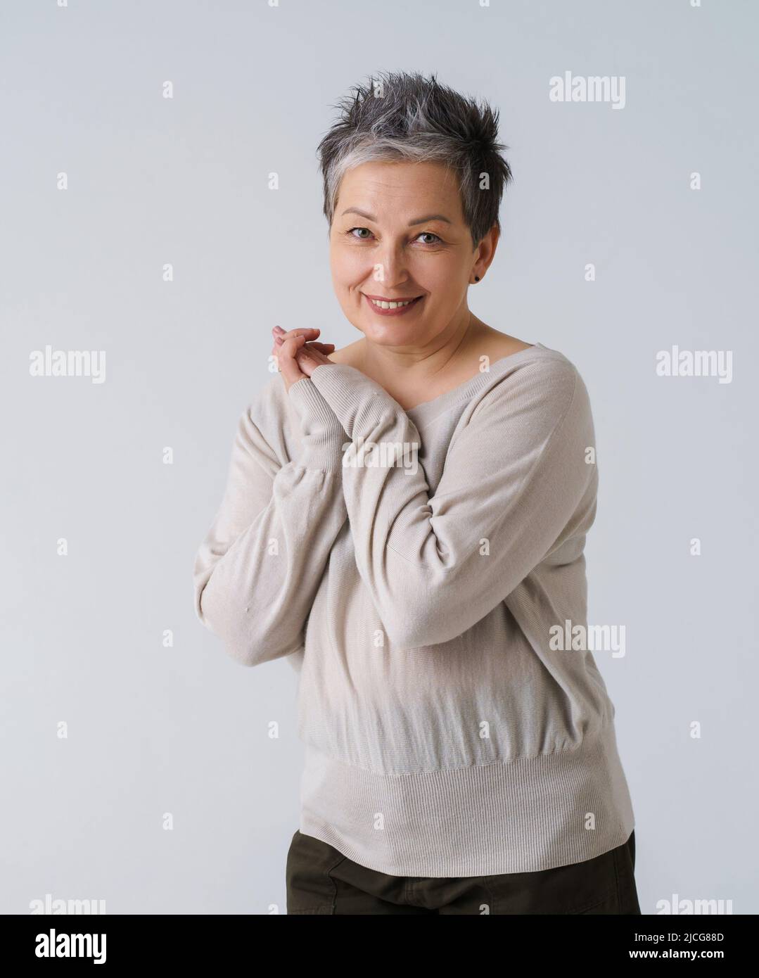 Reife zarte graue Haare Frau in 50s europäischen Ethnie posiert Blick auf Kamera mit gefalteten Händen tragen cremefarbene Bluse isoliert auf weiß. Lächelnde Frau Gesundheit, alternde Schönheit, Kosmetik-Konzept. Stockfoto