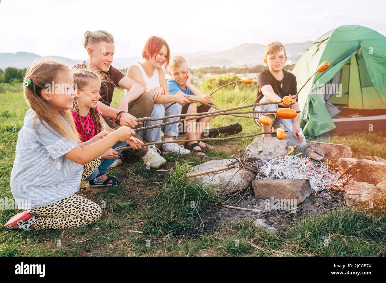 Sechs Kindergruppen Jungen und Mädchen lachten fröhlich und gerösteten Würstchen auf Stöcken über einer Lagerfeuerflamme in der Nähe des grünen Zeltes. Aktive Zeit im Freien Stockfoto