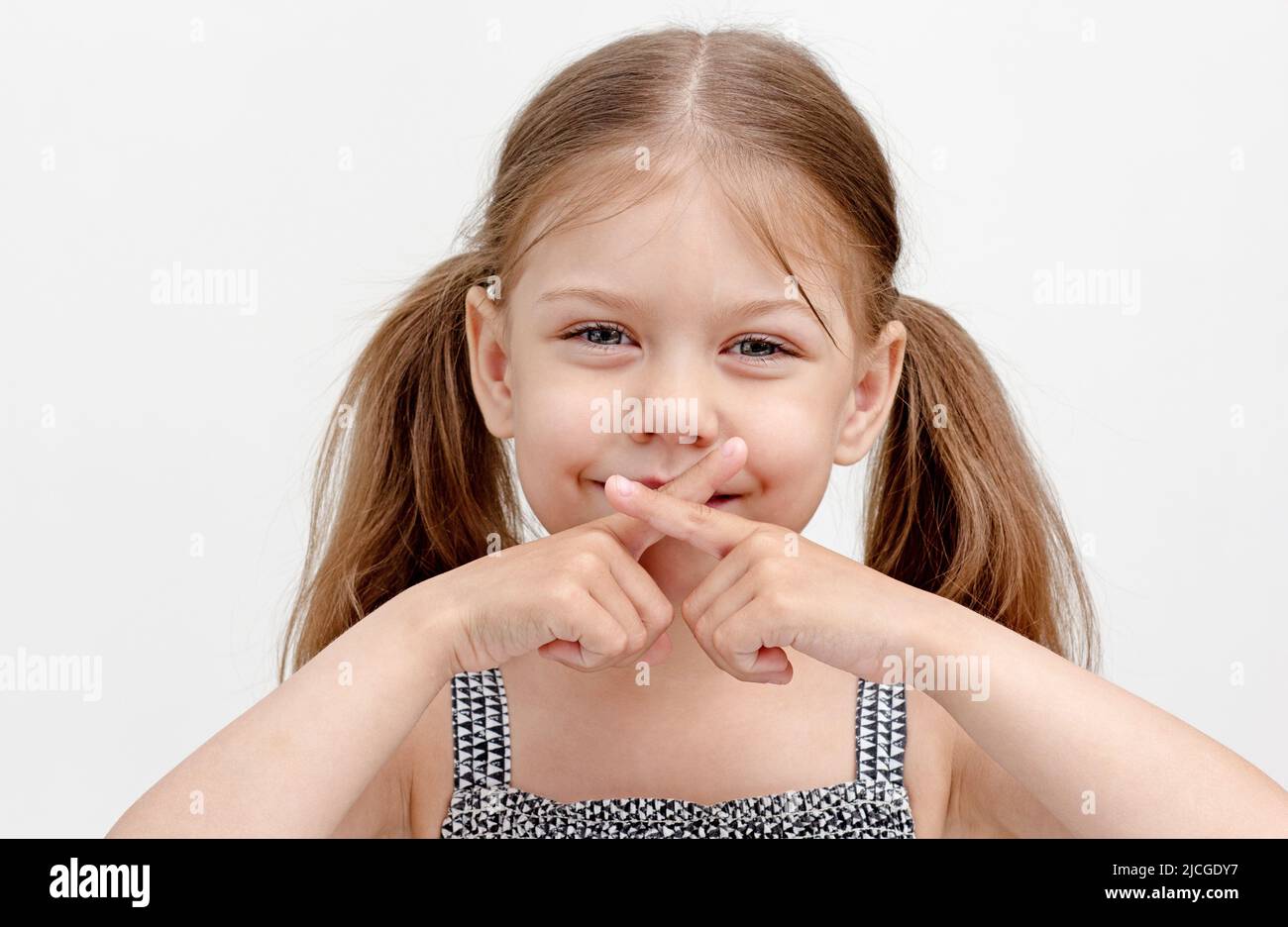 Kaukasisches lächelndes kleines Mädchen von 6 Jahren, das gekreuzte Finger auf dem Mund hält und Schwierigkeiten beim Sprechen zeigt, da es zu Unfähigkeit führt, die Muskeln zu kontrollieren U Stockfoto