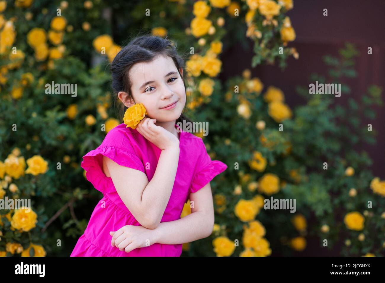 Stilvolle kleine Mädchen 6-7 Jahre alt riechende gelbe Rose Blume über blühenden Büschen im Garten im Freien Nahaufnahme. Blick auf die Kamera. Frühling. Niedlich Stockfoto