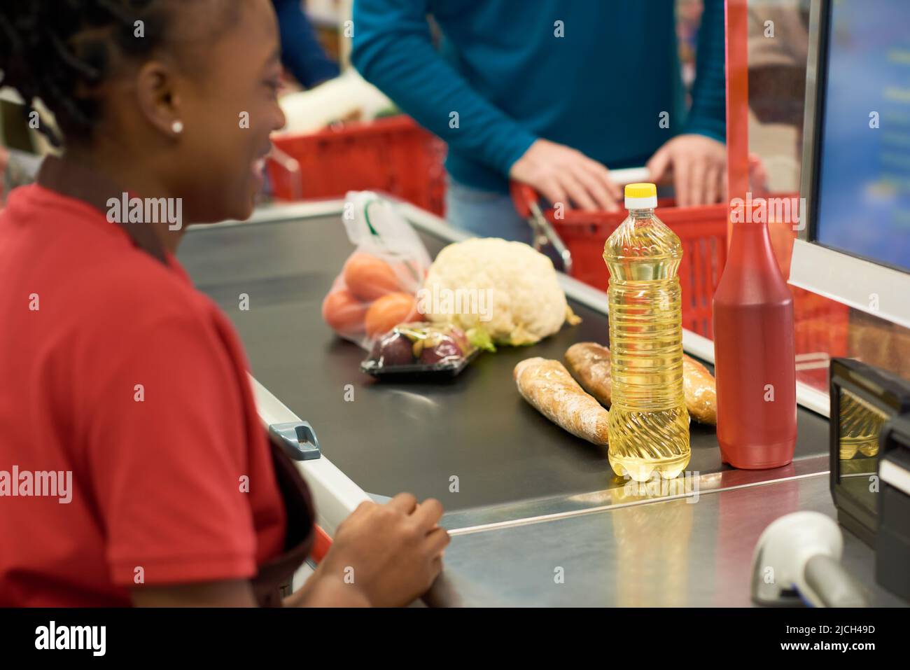 Ketchup, Sonnenblumenöl, Brot und andere frische Lebensmittel an der Kasse zwischen Verkäuferin in Uniform und Käufer mit Wagen Stockfoto