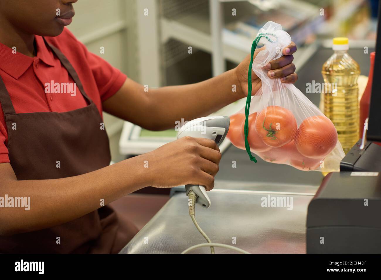 Junge schwarze Frau in rotem Hemd und brauner Schürze scannt frische Tomaten in Zellophanbeutel über den Kassenschalter im Supermarkt Stockfoto