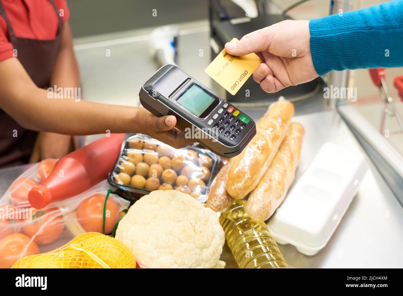 Männliche Verbraucher, die für Lebensmittelprodukte per Kreditkarte bezahlen, während sie sie über das Zahlungsterminal in der Hand der Verkäuferin am Schalter halten Stockfoto