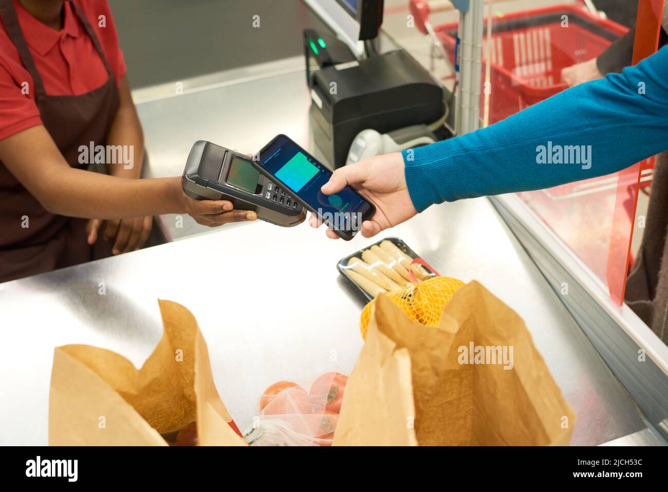 Hand des Käufers mit Smartphone und die des Verkäufers mit Zahlungsterminal über dem Ladentisch mit verpackten Lebensmitteln im Supermarkt Stockfoto