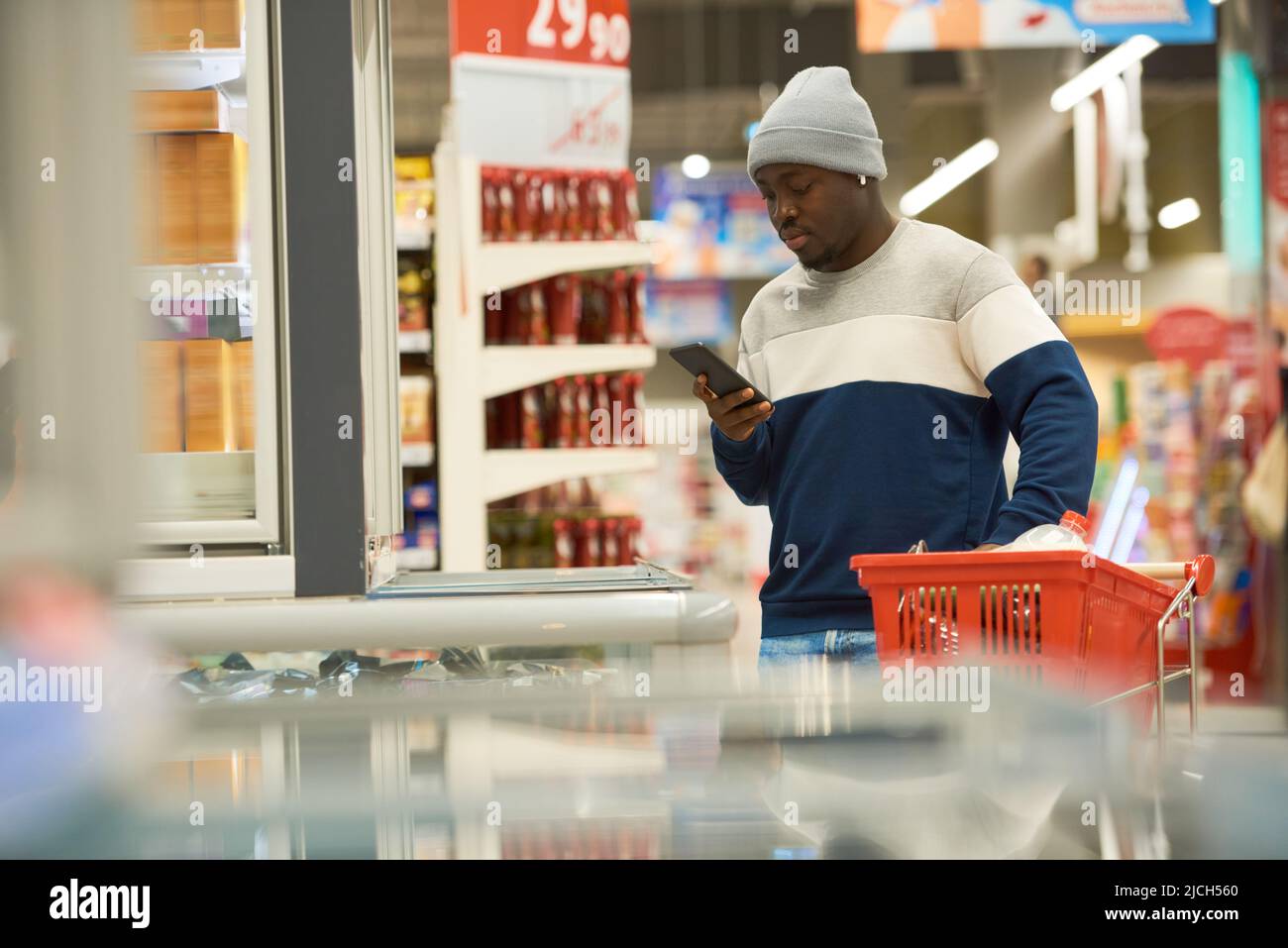 Junger Zoomer in stilvoller Casualwear scrollt mit dem Smartphone, während er in einer der Supermarktabteilungen Lebensmittel auswählt Stockfoto