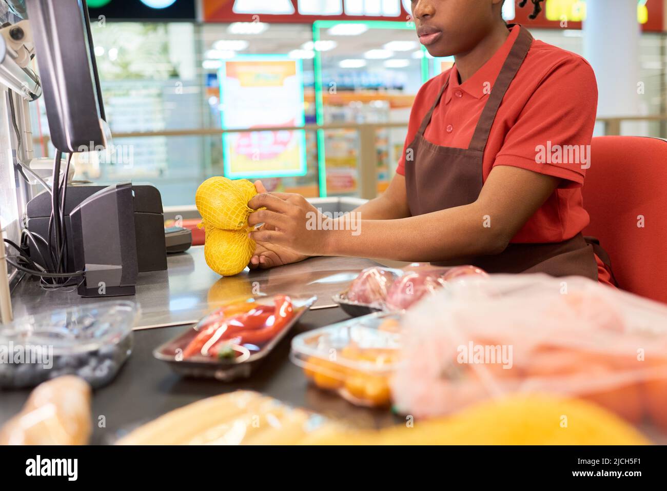Junge afroamerikanische Verkäuferin, die Zitronen in der Tasche wiegt und sie und andere Lebensmittel scannt, während sie an der Theke sitzt Stockfoto