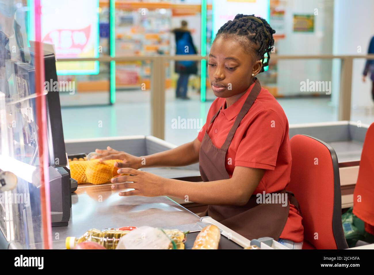 Junge schwarze Frau in Uniform scannt Lebensmittel und legt sie auf andere Seite der Theke, während sie an der Kasse im Supermarkt sitzt Stockfoto