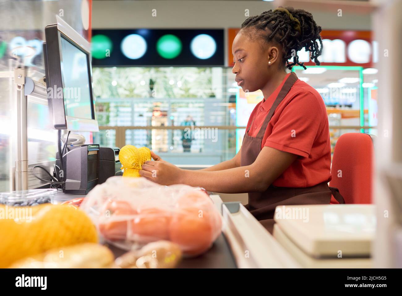 Junge Verkäuferin im Geschäft scannt in Uniform frische Zitronen in Beutel, während sie an der Theke sitzt und Kunden im Supermarkt bedient Stockfoto