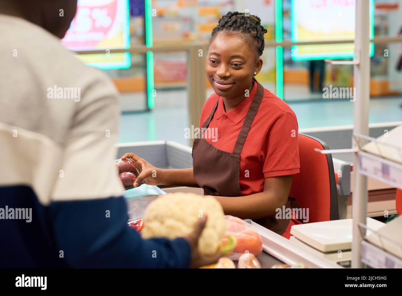 Glückliche junge schwarze Frau in Uniform, die an der Theke sitzt und Lebensmittel scannt, während sie männliche Kunden im Supermarkt bedient Stockfoto