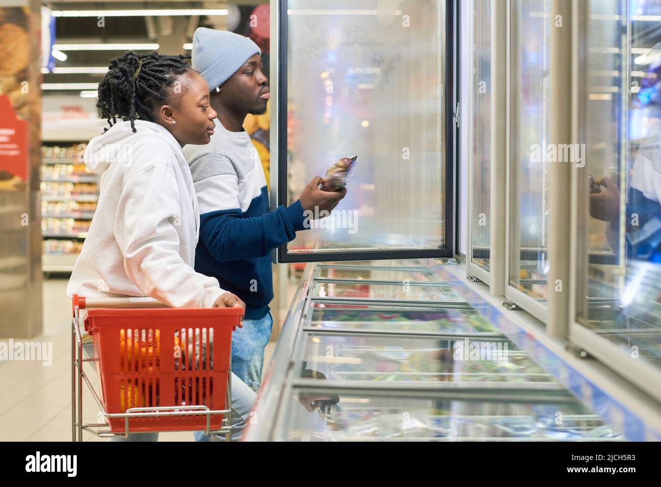 Junges Paar mit Einkaufswagen, das im Supermarkt Tiefkühlprodukte auswählt, während ein Mann eine Obstpackung aus dem Kühlschrank nimmt Stockfoto