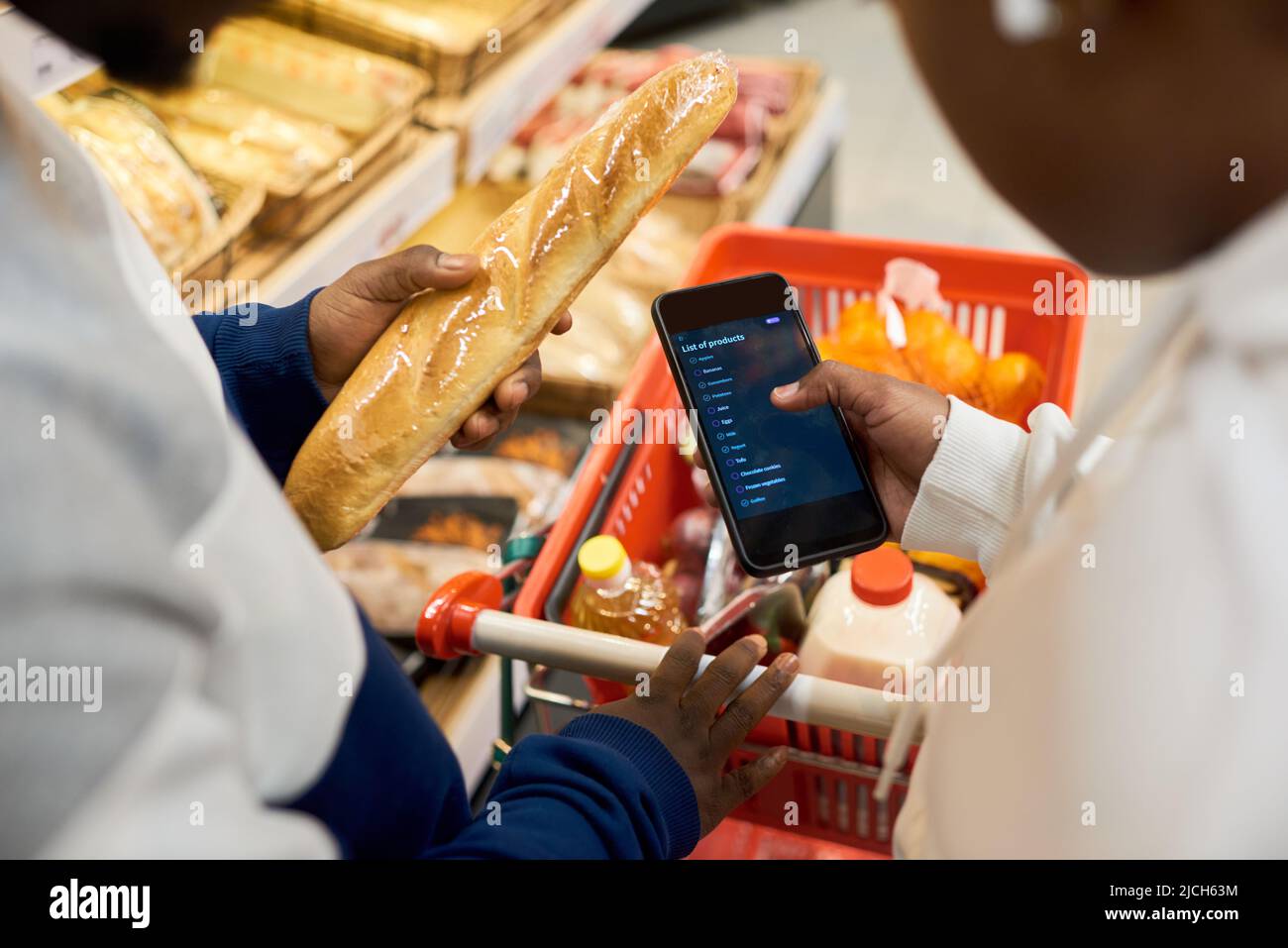 Einkaufsliste auf dem Bildschirm des Smartphones, die von einer jungen afroamerikanischen Frau über dem Wagen mit frischen Lebensmitteln aus der Lebensmittelabteilung gehalten wird Stockfoto