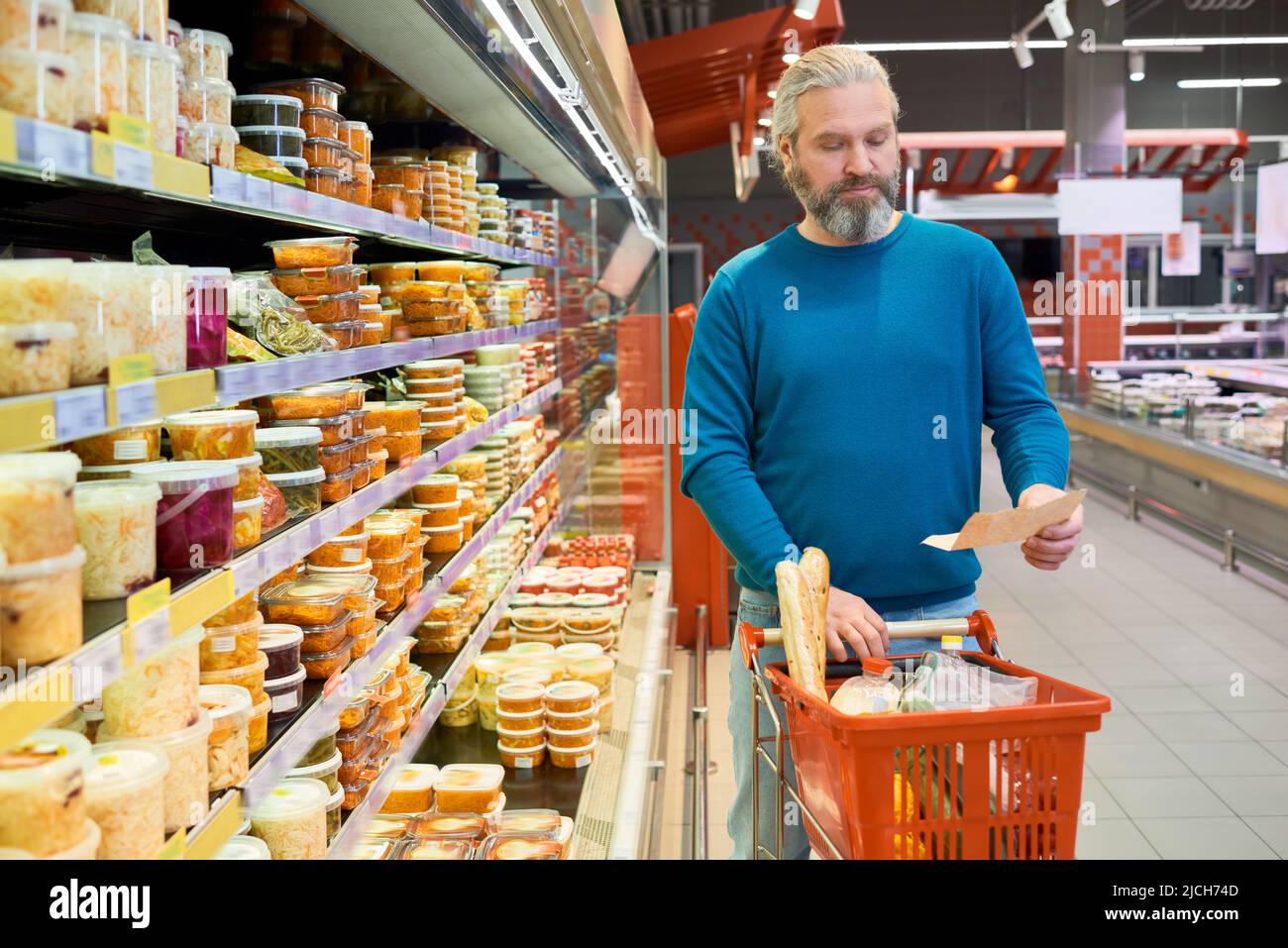 Moderner reifer Mann in Casualwear, der auf die Einkaufsliste schaut, während er sich zwischen den Displays entlang bewegt und den Wagen mit Lebensmittelprodukten schiebt Stockfoto