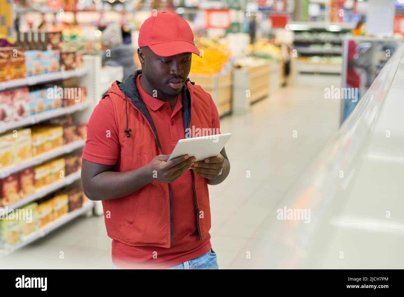 Junger männlicher Arbeiter des Supermarkts, der beim Überprüfen der Preise von Lebensmittelprodukten in der Lebensmittelabteilung den Bildschirm der digitalen Tablette betrachtet Stockfoto
