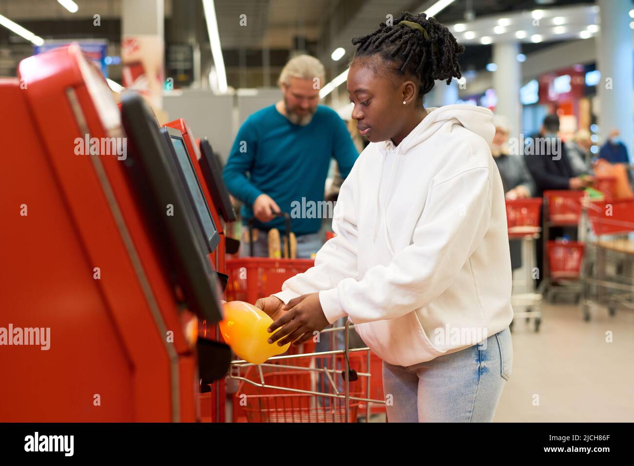 Junge Verbraucherinnen scannen eine Flasche Saft, während sie das Self-Service-Terminal gegen reife männliche Kunden im modernen Supermarkt nutzen Stockfoto
