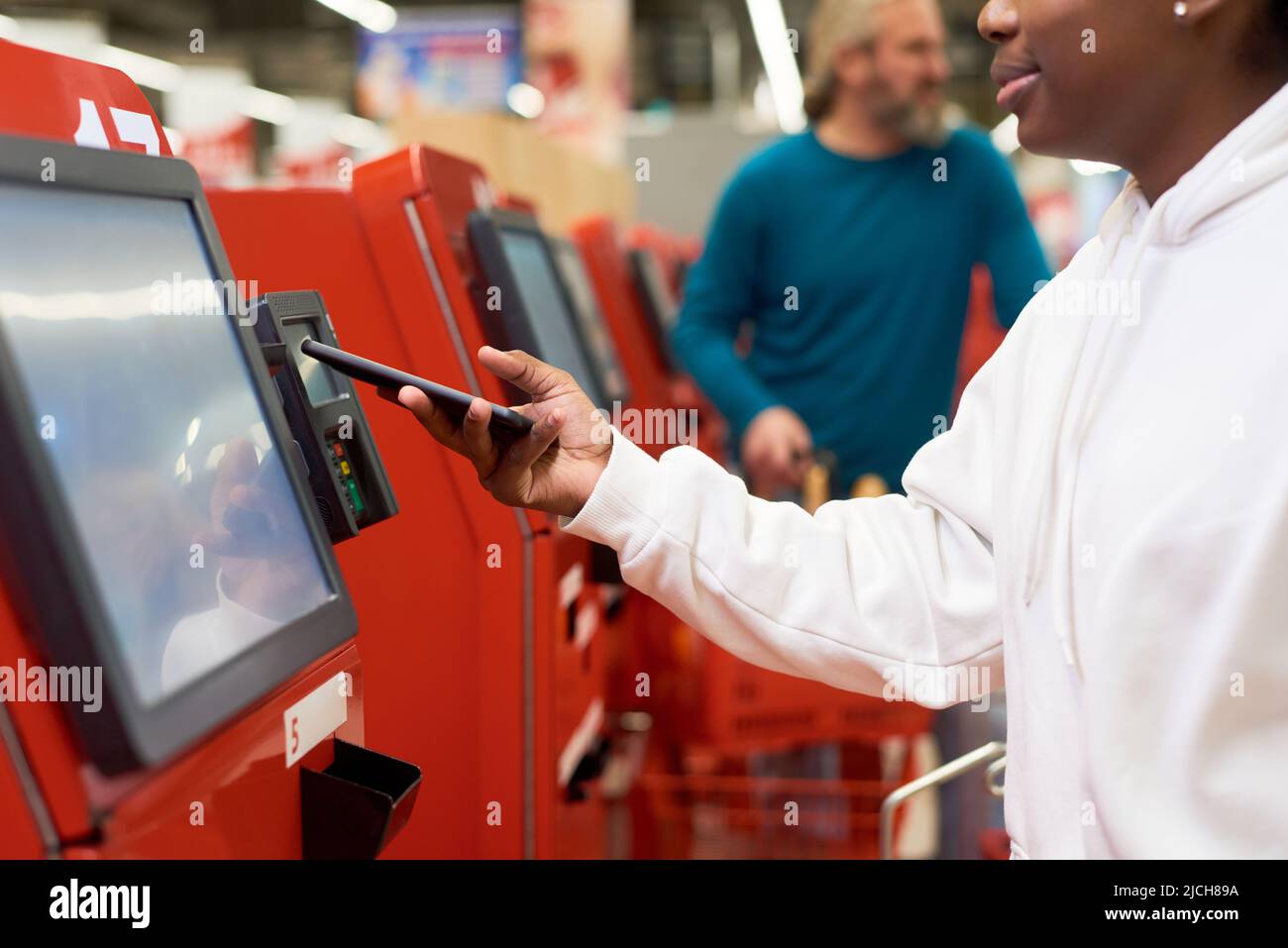 Junge schwarze Frau mit Smartphone, die kontaktlos per Terminal für Self-Service bezahlt, während sie für Lebensmittel im Supermarkt bezahlt Stockfoto