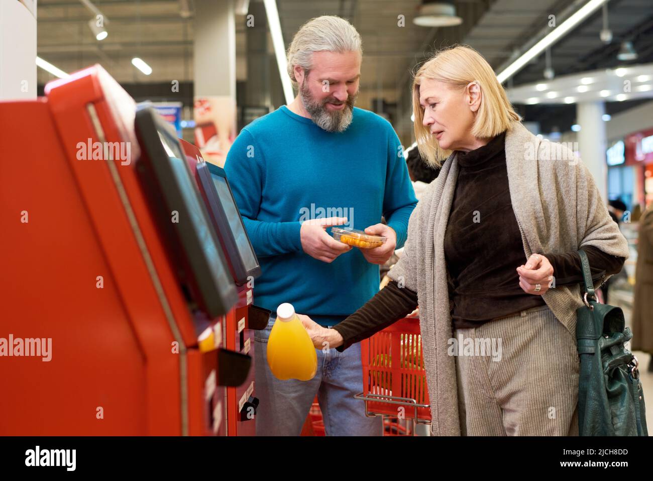Reife blonde Verbraucherin scannt Flasche Orangensaft im Terminal der Selbstbedienung, während sie Lebensmittel im Supermarkt kauft Stockfoto