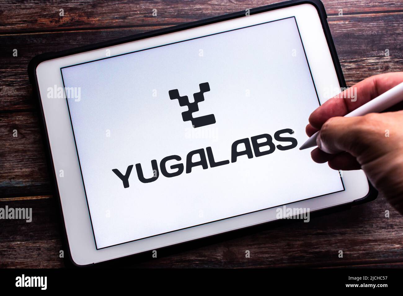 Das Logo von Yuga Labs (der Muttergesellschaft des Bored Ape Yacht Club) auf einem Tablet auf einem Holztisch. Handgriff mit kabellosem Stylus Stockfoto