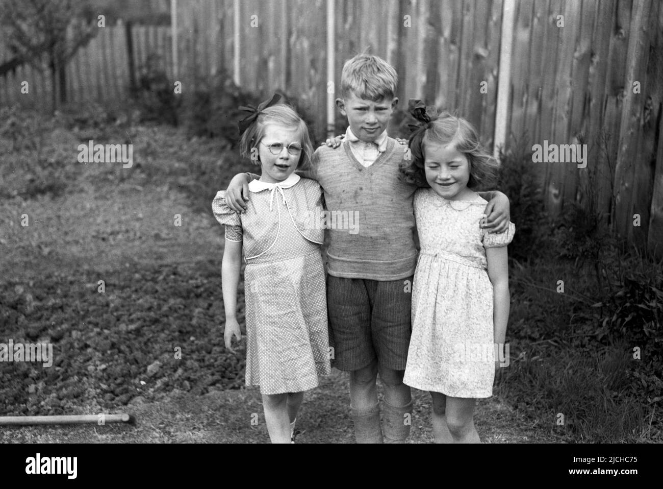 1950s, historisch, draußen im Hintergarten, ein kleiner Junge, der mit seinen Armen um seine beiden jüngeren Schwestern steht, eine lächelnd, mit geschlossenen Augen, ..Schau nicht hin! England, Großbritannien. Stockfoto