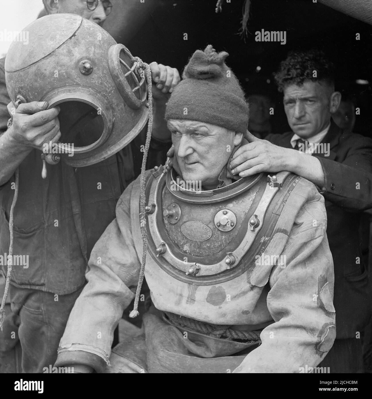 1950s, historisch, ein Tiefseetaucher in der Ausrüstung des Tages, sitzend, mit seinem schweren Messingtauchhelm auf den Kopf gesetzt, im Hafen von London, England, Großbritannien. Stockfoto