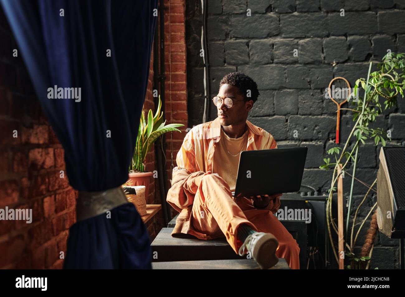 Junger, ernsthafter afroamerikanischer Mann mit Laptop, der an der Fensterbank sitzt und durch das Fenster schaut, während er sich auf die Arbeit konzentriert Stockfoto