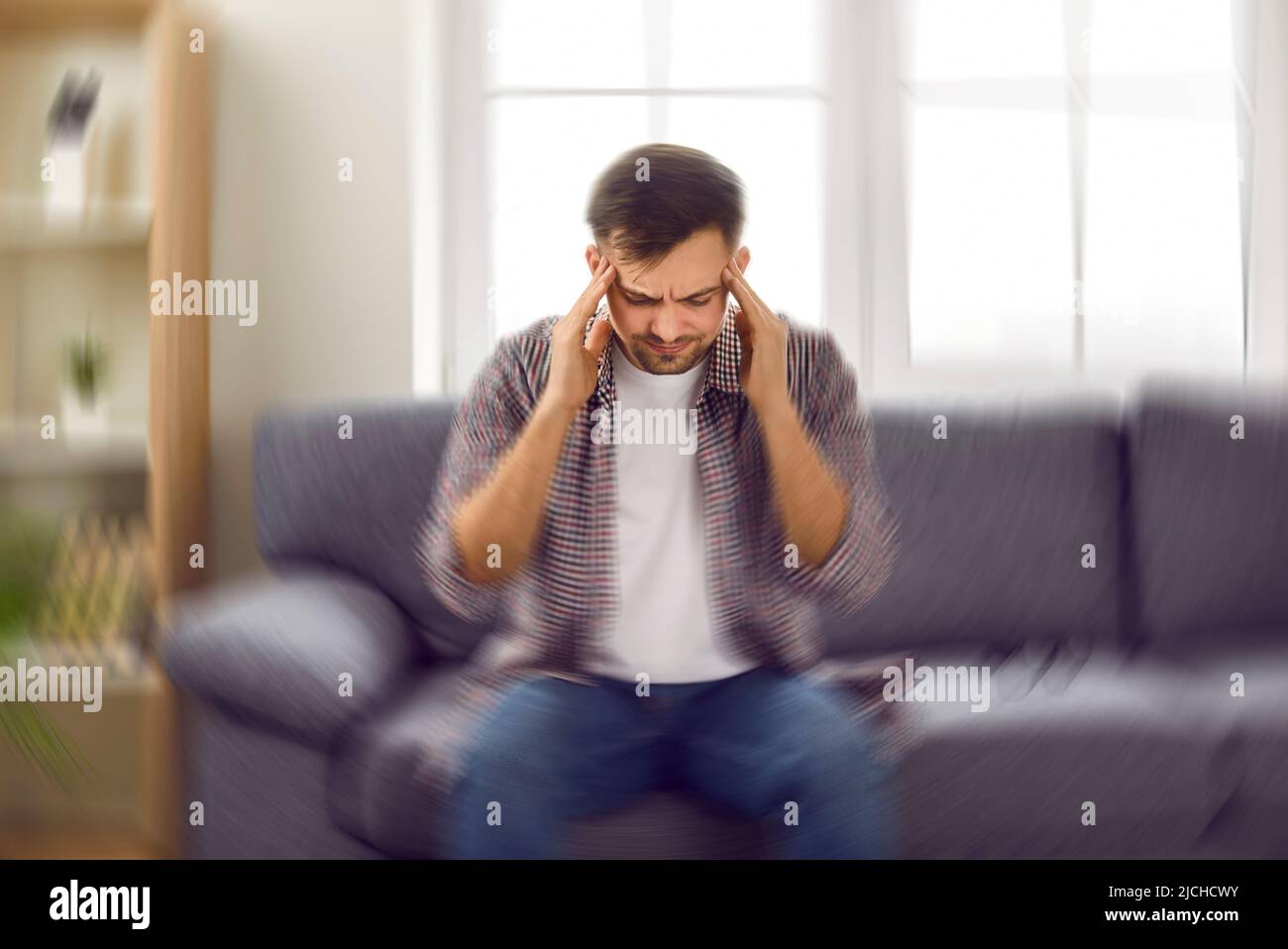 Ein junger Mann, der sich schwindlig fühlt und ein schwindeliges Schwindelgefühl hat, sitzt zu Hause auf dem Sofa Stockfoto