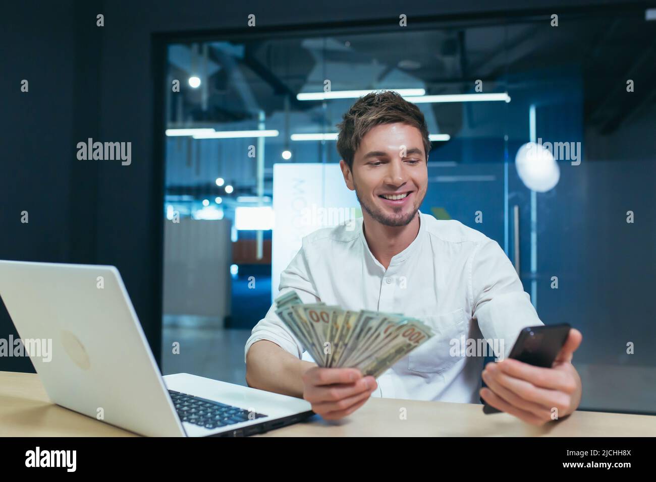 Der Mann arbeitet im Büro, der Geschäftsmann hält Dollar Bargeld freut sich und lächelt schaut auf den Telefonbildschirm, freut sich über den Sieg Stockfoto