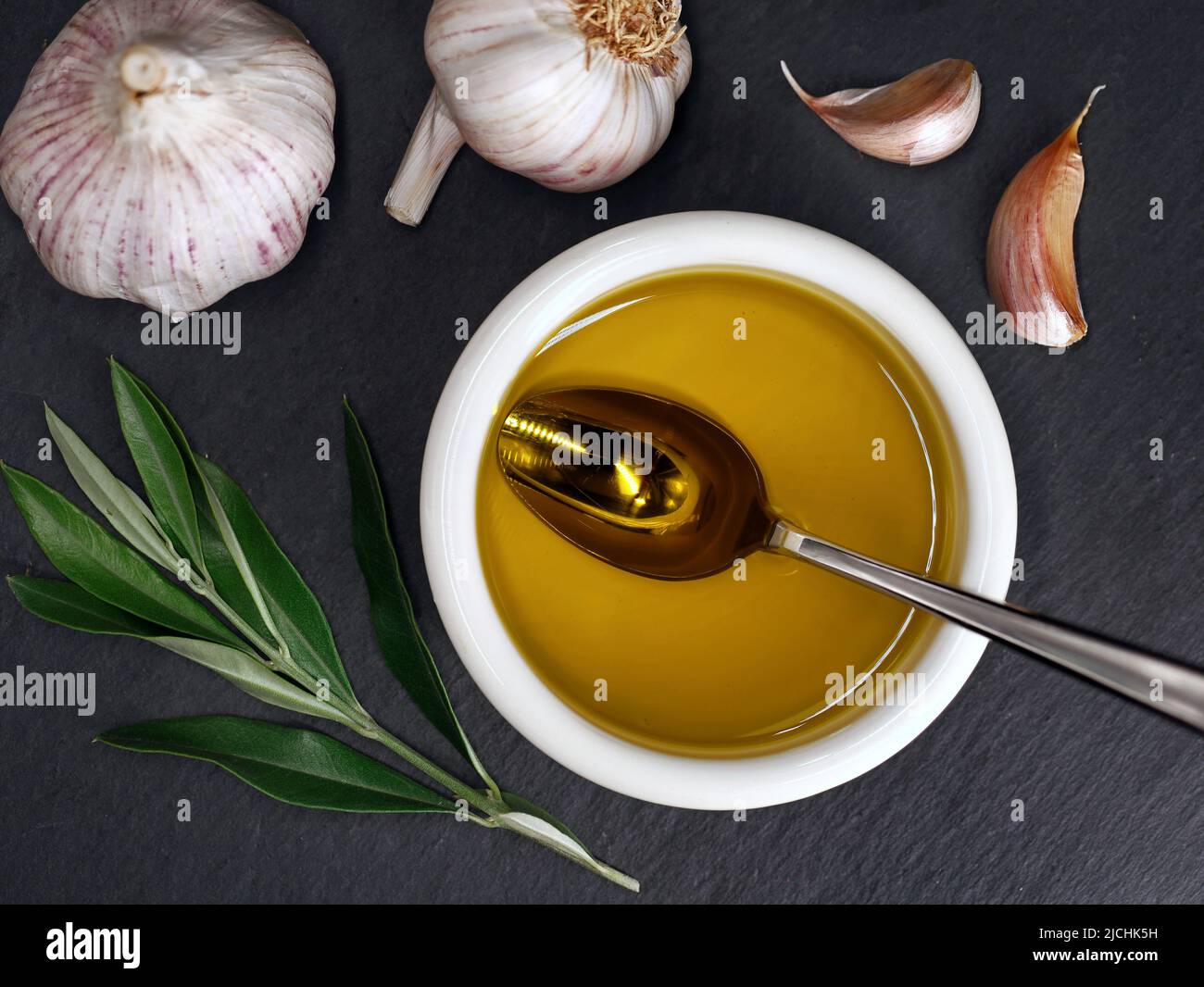 Draufsicht auf eine Schüssel mit nativem Olivenöl auf Schieferteller mit Knoblauch und Olivenbaum Zweig Stockfoto